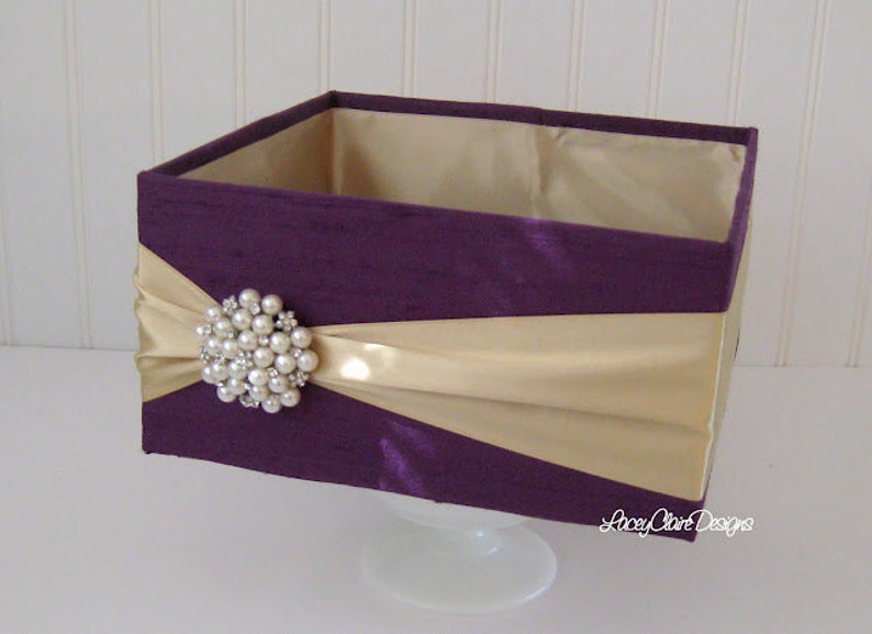 May include: A square box with a purple fabric exterior and a gold satin ribbon. The box is decorated with a large pearl brooch.