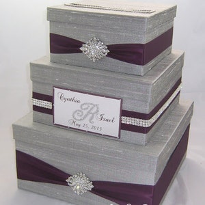 May include: Three-tiered gray gift boxes with purple ribbon and rhinestone embellishments. The boxes have a slot on top and a decorative label with the names "Cynthia R. Isael" and the date "May 25, 2013".