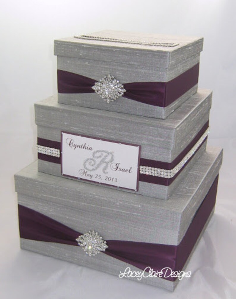 May include: Three-tiered gray gift boxes with purple ribbon and rhinestone embellishments. The boxes have a slot on top and a decorative label with the names "Cynthia R. Isael" and the date "May 25, 2013".