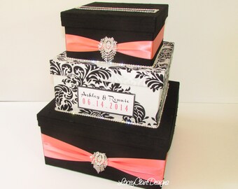May include: Three-tiered wedding card box with black and white damask pattern on the middle tier. The top and bottom tiers are black with a coral satin ribbon and a rhinestone embellishment. The middle tier has a white label with the text "Ashley & Ronnie 06.14.2014".