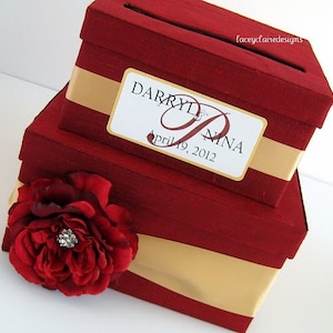 May include: Two-tiered red wedding card box with a gold ribbon and a white label with the names "Darryl & Nina" and the date "April 19, 2012". A red rose with a rhinestone embellishment is on the front of the box.