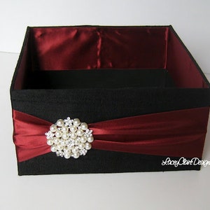 May include: A black gift box with a burgundy satin lining and a burgundy satin ribbon tied around it. The ribbon is adorned with a large pearl and rhinestone brooch. The box is labeled "Lucy Claire Designs".