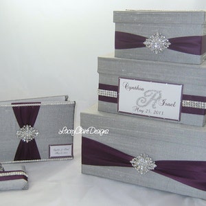 May include: A set of silver-colored wedding accessories, including a guest book, pen holder, and three decorative boxes. Each item features a deep purple ribbon and a sparkling rhinestone brooch. The boxes are different sizes.