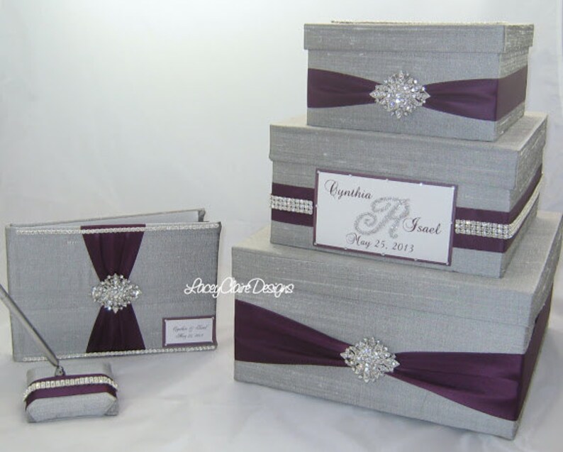 May include: A set of silver-colored wedding accessories, including a guest book, pen holder, and three decorative boxes. Each item features a deep purple ribbon and a sparkling rhinestone brooch. The boxes are different sizes.