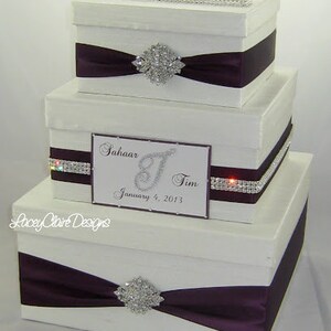 May include: Three-tiered white gift box set with dark purple satin ribbon and rhinestone accents. Each box has a decorative rhinestone brooch. The middle box has a card with the names "Sahaar & Tim" and the date "January 4, 2013".