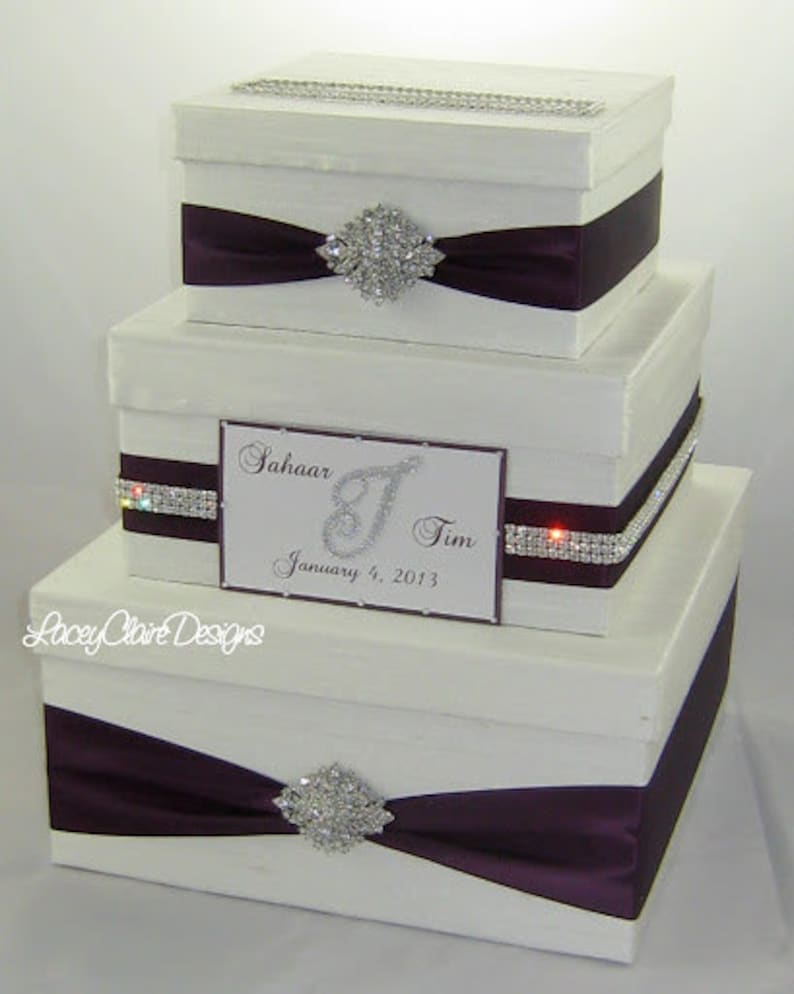 May include: Three-tiered white gift box set with dark purple satin ribbon and rhinestone accents. Each box has a decorative rhinestone brooch. The middle box has a card with the names "Sahaar & Tim" and the date "January 4, 2013".