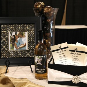 May include: A black picture frame with a gold patterned border, a bottle of wine with a personalized label, and a black and white box with a white satin ribbon and a pearl brooch. The box contains a stack of cards with the text "Menu of Events" printed on them.