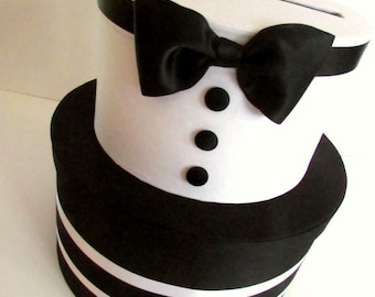 May include: A decorative box in the style of a tuxedo, with a white body and black accents. It has a black bow tie, three black buttons, and a slot. The base is striped black and white.