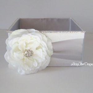 Pode incluir: Uma caixa quadrada branca com uma flor decorativa na frente. A caixa tem uma borda de glitter prateada e o texto "Lacey Olive Designs" no canto inferior direito.