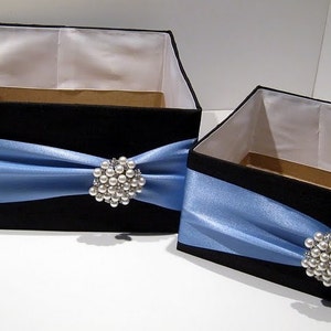 May include: Two black gift boxes with blue satin ribbon and pearl brooches. The boxes are of different sizes.