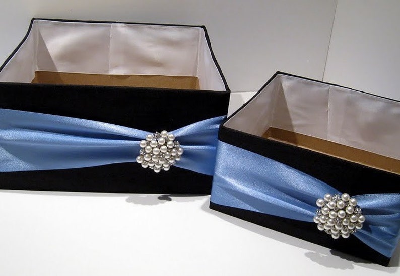May include: Two black gift boxes with blue satin ribbon and pearl brooches. The boxes are of different sizes.
