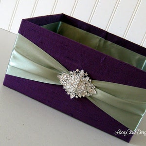 Pode incluir: Uma caixa de veludo roxo com uma fita de cetim verde-sálvia e um broche de strass prateado. A caixa é perfeita para um casamento ou outra ocasião especial.
