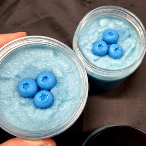 May include: Two clear plastic jars filled with light blue, shimmering, textured material. Each jar is topped with three bright blue, blueberry-shaped decorations. The jars are open, revealing the contents. The background is a dark, neutral color.