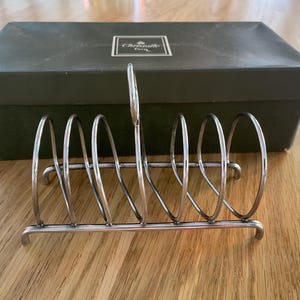 Könnte beinhalten: Silberfarbener Metall-Toastständer mit sechs gebogenen Schlitzen, der auf einer rechteckigen Basis ruht. Eine dunkelgrüne Schachtel befindet sich hinter dem Ständer. Die Schachtel hat ein weißes Etikett mit schwarzem Text.