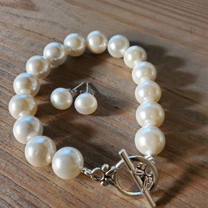 Puede incluir: Un brazalete de perlas color crema y pendientes a juego. El brazalete presenta perlas grandes y redondas con un cierre plateado. Los pendientes son peque&ntilde;as perlas redondas con postes plateados. La joyer&iacute;a se exhibe sobre una superficie de madera.