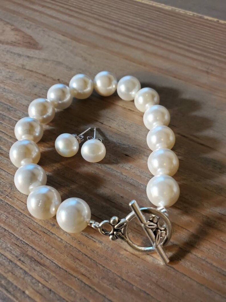 Puede incluir: Un brazalete de perlas color crema y pendientes a juego. El brazalete presenta perlas grandes y redondas con un cierre plateado. Los pendientes son peque&ntilde;as perlas redondas con postes plateados. La joyer&iacute;a se exhibe sobre una superficie de madera.