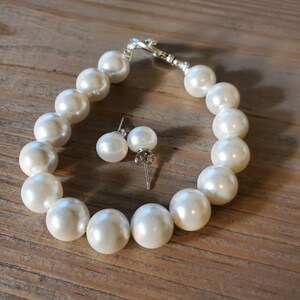 Puede incluir: Un juego de pulsera de perlas color crema y pendientes a juego. La pulsera presenta perlas grandes y redondas ensartadas con un cierre plateado. Los pendientes tienen perlas similares en postes plateados. El conjunto de joyas se exhibe sobre una superficie de madera.