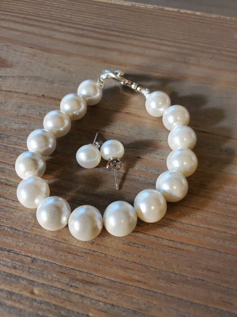 Puede incluir: Un juego de pulsera de perlas color crema y pendientes a juego. La pulsera presenta perlas grandes y redondas ensartadas con un cierre plateado. Los pendientes tienen perlas similares en postes plateados. El conjunto de joyas se exhibe sobre una superficie de madera.