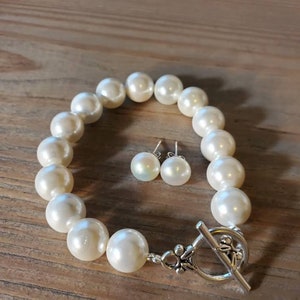 Puede incluir: Un brazalete de perlas color crema y pendientes a juego. El brazalete presenta perlas grandes y redondas ensartadas con un cierre plateado. Los pendientes son peque&ntilde;as perlas redondas. Las joyas se exhiben sobre una superficie de madera.