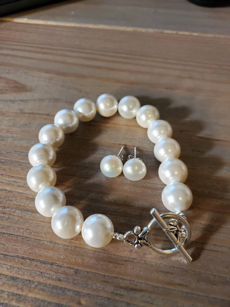 Puede incluir: Un brazalete de perlas color crema y pendientes a juego. El brazalete presenta perlas grandes y redondas ensartadas con un cierre plateado. Los pendientes son peque&ntilde;as perlas redondas. Las joyas se exhiben sobre una superficie de madera.