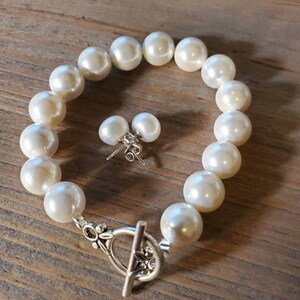 Puede incluir: Un brazalete de perlas color crema y pendientes a juego. El brazalete presenta perlas grandes y redondas ensartadas con un cierre plateado. Los pendientes son peque&ntilde;as perlas redondas con postes plateados.