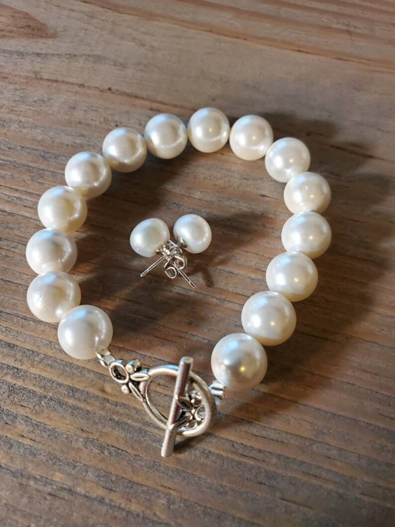 Puede incluir: Un brazalete de perlas color crema y pendientes a juego. El brazalete presenta perlas grandes y redondas ensartadas con un cierre plateado. Los pendientes son peque&ntilde;as perlas redondas con postes plateados.