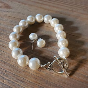 Puede incluir: Un brazalete de perlas color crema y pendientes a juego. El brazalete presenta grandes perlas redondas ensartadas con un cierre plateado. Los pendientes tienen una sola perla cada uno, montados en postes plateados.