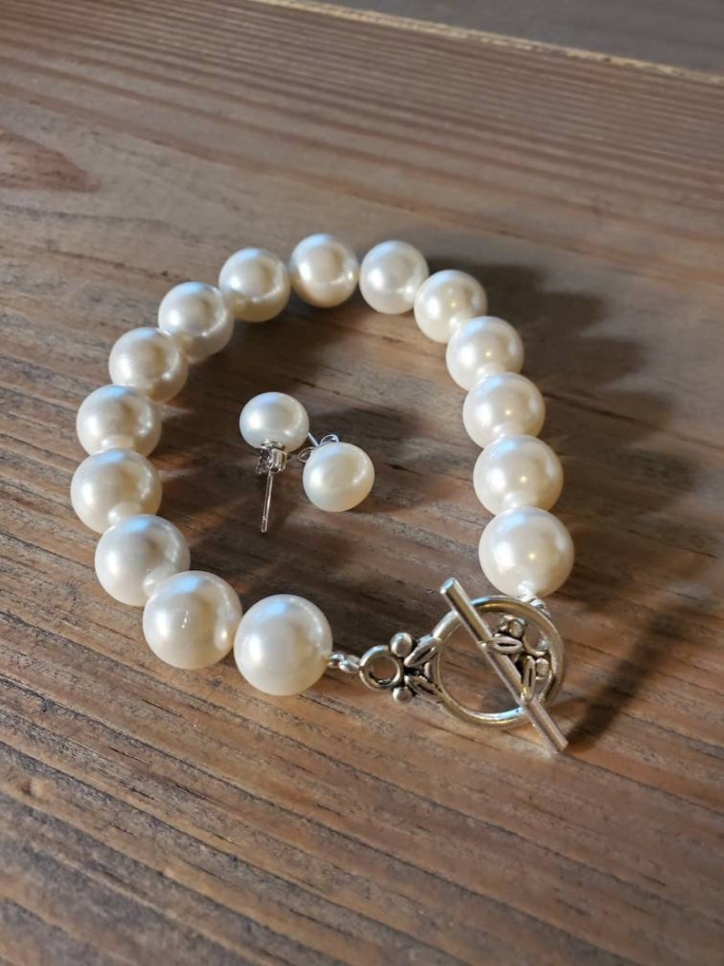 Puede incluir: Un brazalete de perlas color crema y pendientes a juego. El brazalete presenta grandes perlas redondas ensartadas con un cierre plateado. Los pendientes tienen una sola perla cada uno, montados en postes plateados.