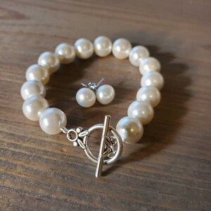 Puede incluir: Un juego de pulsera de perlas color crema y pendientes a juego. La pulsera presenta perlas grandes y redondas unidas con un cierre de palanca plateado. Los pendientes son sencillos, con una sola perla en cada pendiente.