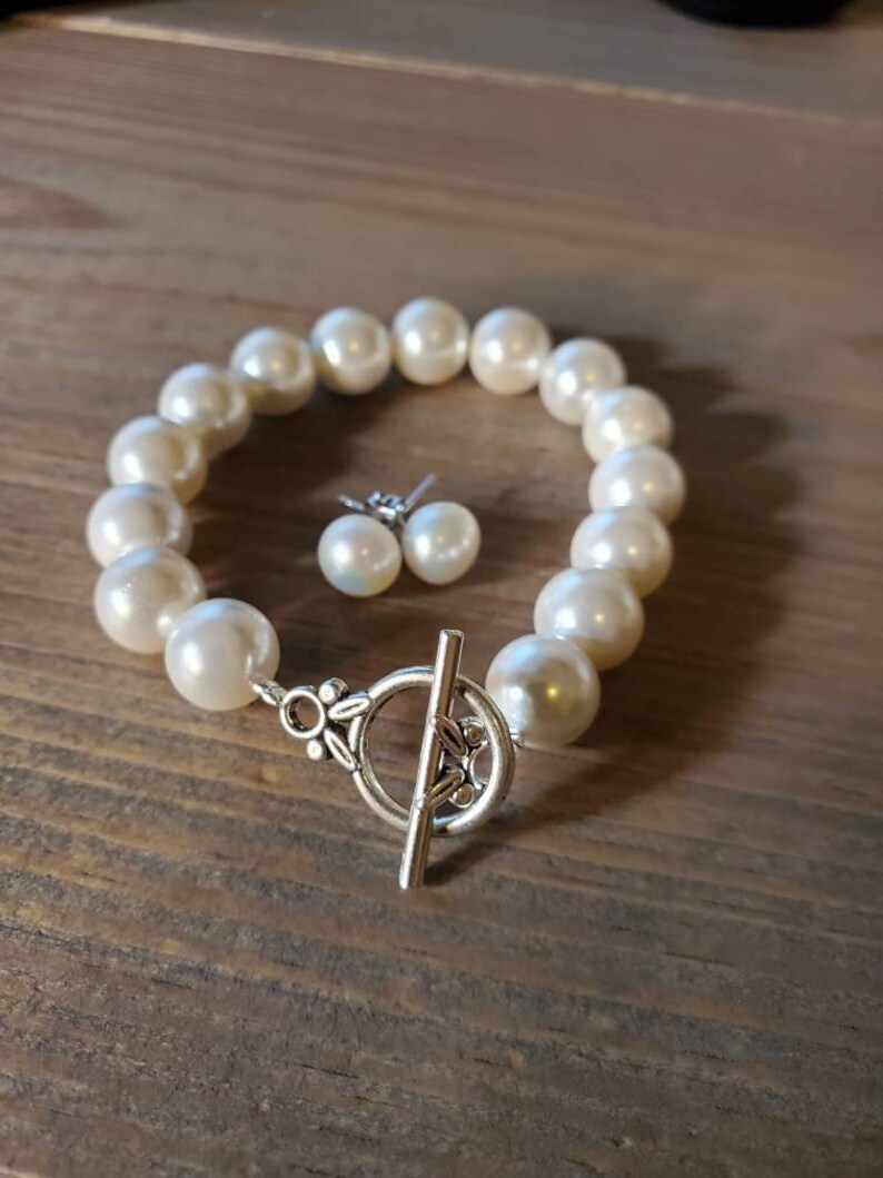 Puede incluir: Un juego de pulsera de perlas color crema y pendientes a juego. La pulsera presenta perlas grandes y redondas unidas con un cierre de palanca plateado. Los pendientes son sencillos, con una sola perla en cada pendiente.