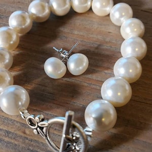 Puede incluir: Una pulsera de perlas color crema con un cierre plateado y pendientes de perlas a juego. La pulsera presenta perlas grandes y redondas. Los pendientes son perlas peque&ntilde;as y redondas con postes plateados. El conjunto de joyas se exhibe sobre una superficie de madera.