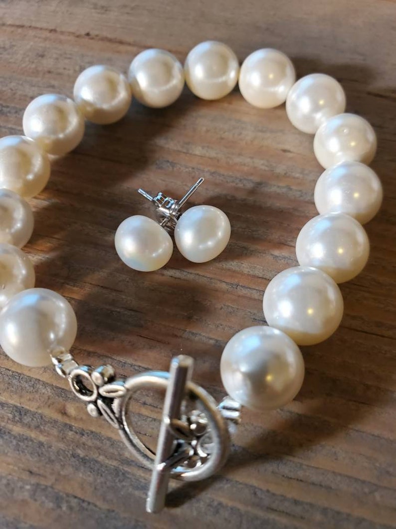 Puede incluir: Una pulsera de perlas color crema con un cierre plateado y pendientes de perlas a juego. La pulsera presenta perlas grandes y redondas. Los pendientes son perlas peque&ntilde;as y redondas con postes plateados. El conjunto de joyas se exhibe sobre una superficie de madera.