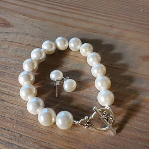 Puede incluir: Un juego de pulsera de perlas color crema y pendientes a juego sobre una superficie de madera. La pulsera presenta perlas grandes y redondas y un cierre de palanca plateado. Los pendientes tienen una sola perla cada uno.