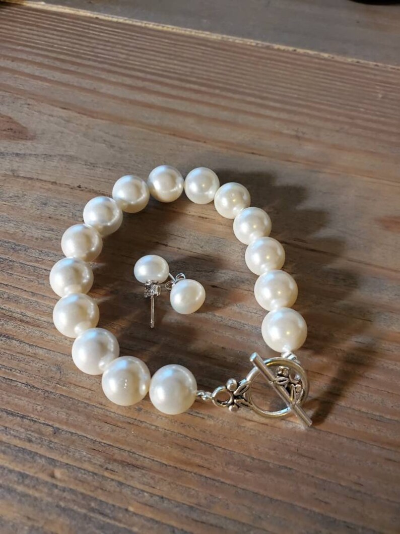 Puede incluir: Un juego de pulsera de perlas color crema y pendientes a juego sobre una superficie de madera. La pulsera presenta perlas grandes y redondas y un cierre de palanca plateado. Los pendientes tienen una sola perla cada uno.