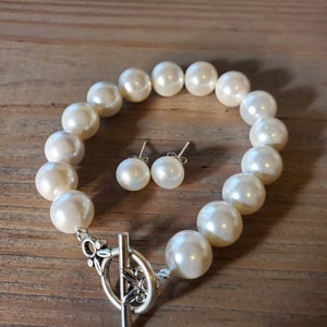 Puede incluir: Un brazalete de perlas color crema y pendientes a juego. El brazalete presenta perlas grandes y redondas con un cierre decorativo plateado. Los pendientes tienen una sola perla cada uno. La joyer&iacute;a se exhibe sobre una superficie de madera.