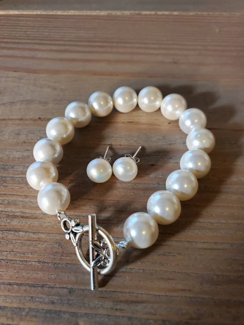 Puede incluir: Un brazalete de perlas color crema y pendientes a juego. El brazalete presenta perlas grandes y redondas con un cierre decorativo plateado. Los pendientes tienen una sola perla cada uno. La joyer&iacute;a se exhibe sobre una superficie de madera.