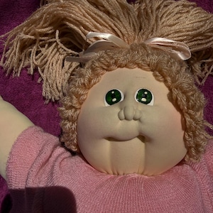 May include: A soft-sculpture doll with a round fabric face, green eyes, and a small nose. The doll has light brown yarn hair styled with a pale pink ribbon bow. It is wearing a short-sleeved, light pink knit shirt with a textured, ribbed pattern.