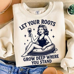 May include: Cream-colored sweatshirt with a graphic of a woman in a swimsuit holding a wine glass, sitting in a pool float. Text reads "LET YOUR ROOTS GROW DEEP WHERE YOU STAND".