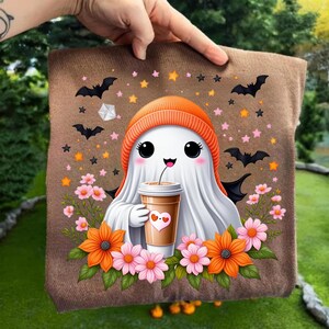 Könnte beinhalten: Ein braunes Halloween-Kissen mit einem niedlichen Geist, der eine orangefarbene Mütze trägt und einen Kaffeebecher mit einem Herz hält. Das Kissen ist mit Fledermäusen, Sternen und Blumen verziert.