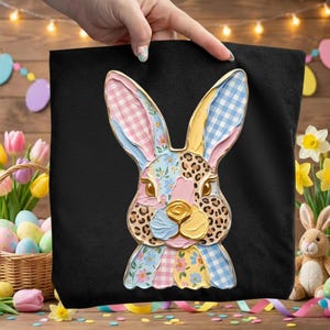 Op de afbeelding: Zwarte tote bag met een kleurrijk paashaasontwerp. De haas heeft een patchworkpatroon met roze, blauwe, gele en luipaardprint accenten. De oren van de haas zijn roze en blauw geruit en het gezicht heeft bloemendetails.