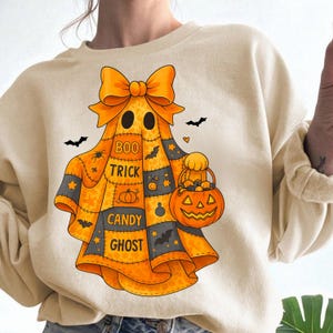 May include: Cream-colored sweatshirt featuring a Halloween ghost design. The ghost is orange and black with a large bow, holding a jack-o'-lantern basket. The words "Boo Trick Candy Ghost" are printed on the ghost. Black bats and a heart are also included.