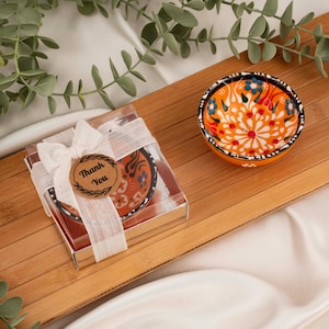 May include: A small, round, orange ceramic bowl with a floral design in blue, red, and white. The bowl is on a bamboo tray. A clear box with a matching bowl is tied with a white ribbon and a "Thank You" tag.