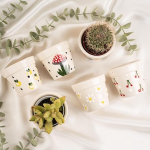 May include: A selection of small, cream ceramic plant pots with hand-painted designs. The pots display illustrations of lemons, cherries, daisies, and a mushroom. One pot contains a cactus, and another holds a succulent. The pots are arranged on a white, textured surface.