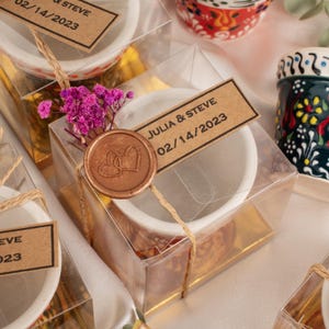 Puede incluir: Pequeños cuencos de cerámica blanca empaquetados en cajas transparentes, atados con hilo y sellados con un sello de cera con dos corazones. Cada caja tiene una etiqueta que dice "JULIA & STEVE 02/14/2023". Una ramita de flores rosas.