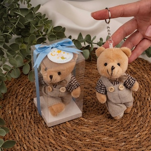 Puede incluir: Un pequeño llavero de oso de peluche marrón con una camisa a cuadros marrón y un peto beige. El oso tiene un llavero de metal adjunto. Otro oso está dentro de una caja transparente con una cinta azul y una etiqueta que dice "Little Prince".