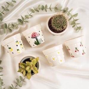 Può includere: Una collezione di piccoli vasi di ceramica color crema con disegni dipinti a mano. I vasi presentano illustrazioni di limoni, ciliegie, margherite e un fungo. Un vaso contiene un cactus e un altro una pianta succulenta.