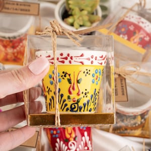 May include: A hand-painted ceramic pot with a yellow base and colourful floral designs. The pot is held in a clear box tied with twine. The pot's rim is white with red accents. Other pots are visible in the background.