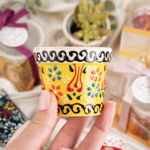 Könnte beinhalten: Ein kleiner, handbemalter Keramiktopf mit leuchtend gelbem Grund. Der Topf hat ein sich wiederholendes Blumenmuster in Rot, Blau und Weiß mit schwarz-weißen Zierleisten. Der Topf wird in einer Hand gehalten, was auf seine geringe Größe schließen lässt, und ist wahrscheinlich für eine Sukkulente.