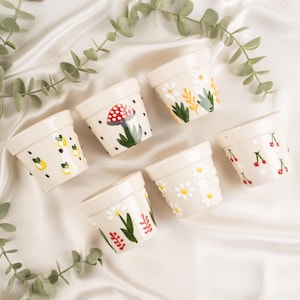 Puede incluir: Seis pequeñas macetas de cerámica blanquecina con diseños pintados a mano. Las macetas presentan ilustraciones de limones, cerezas, margaritas y una seta. Las macetas están dispuestas sobre una superficie blanca y sedosa con follaje verde.