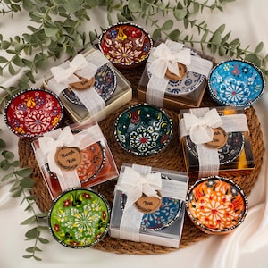Può includere: Una collezione di ciotole in ceramica dipinte a mano, ognuna con motivi floreali unici. Le ciotole sono confezionate in scatole trasparenti, legate con nastri bianchi e piccoli tag "Thank You". Le ciotole sono disposte su un tappetino intrecciato.